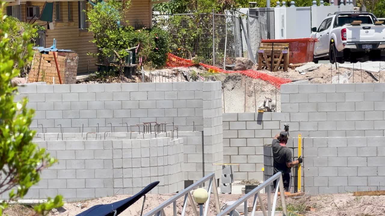 Worker uses spirit level on concrete block wall at residential construction site, midday sunlight