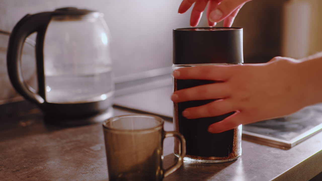 Close up of coffee container placed on kitchen counter beside electric kettle, hand visible above preparing to open container and scoop granules into cup, warm morning light filling cozy atmosphere