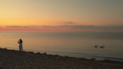 Drone-follows-woman-fixing-hair-walking-along-beach-coast,-sunset-glow-in-sky-and-water