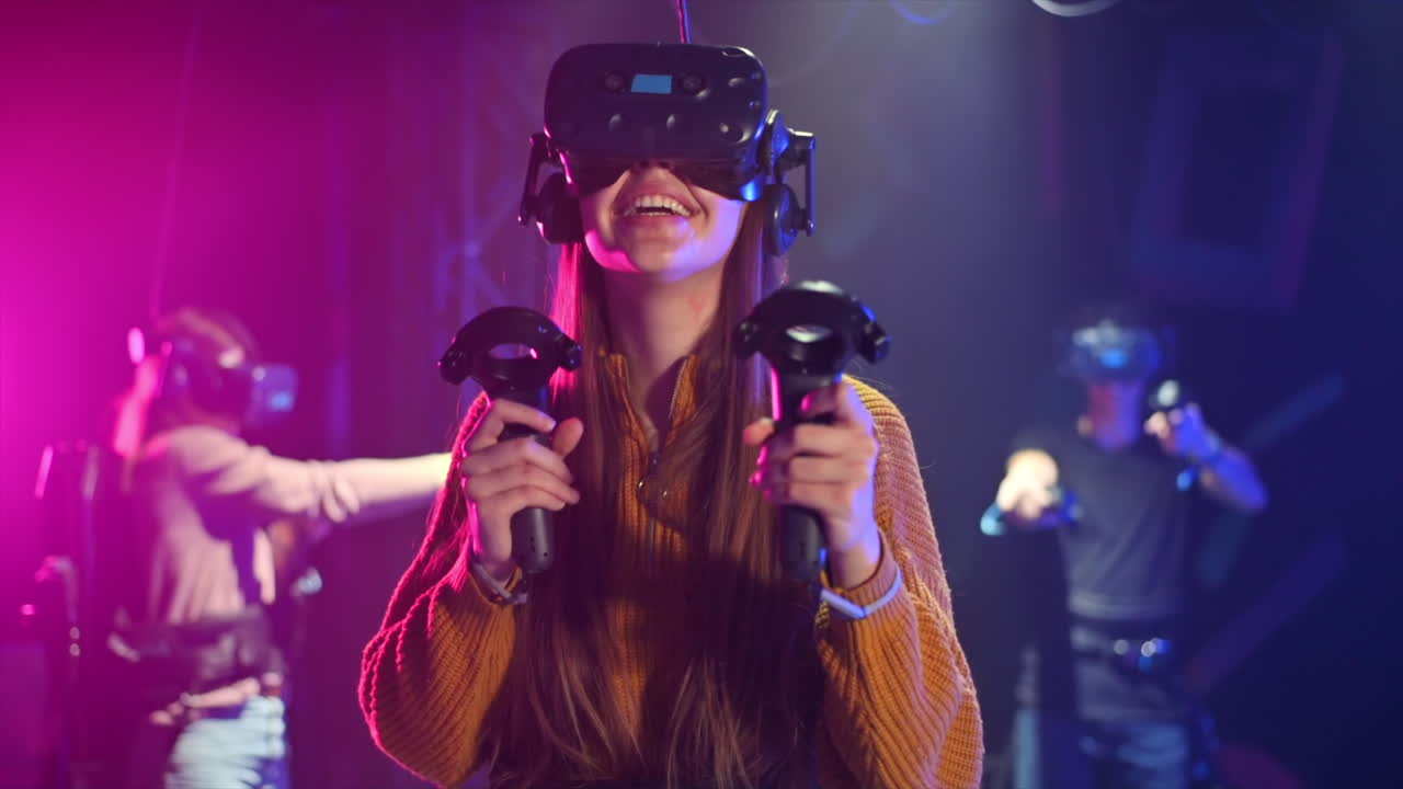 Una bambina felice e interessata gioca su un tapis roulant VR con casco e controller. Altre persone giocano nelle vicinanze. Slow motion