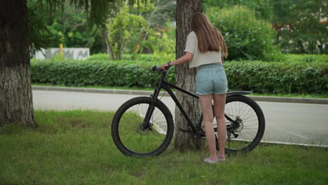 woman in jean shorts approaches bicycle leaning against tree in serene park setting, reaching to remove stand, surrounded by lush green background of trees and paved path