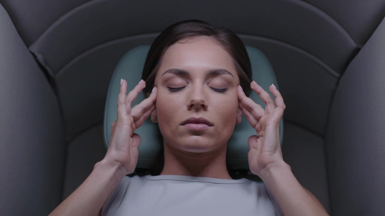 Reclining in capsule, woman in sleeveless top massaging temples with fingers on headrest, relaxing