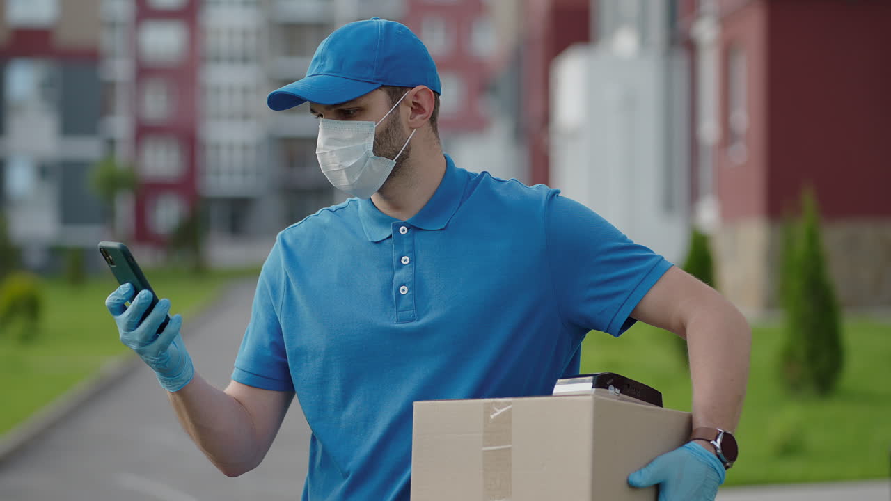 The male deliveryman in a cap and a protective mask and gloves goes with a box in his hands and carries a parcel to the customer. Delivering online home orders.