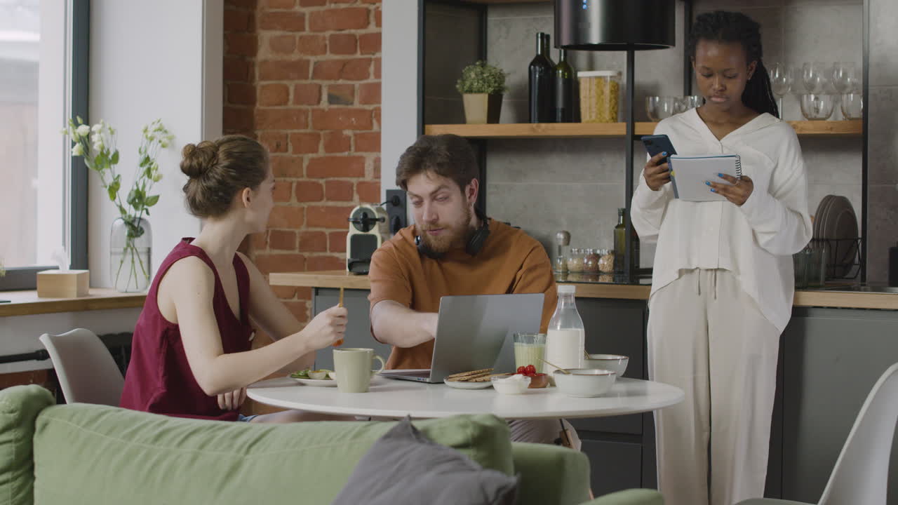 세 명의 룸메이트가 집에서 아침 식사를 하는 동안 컴퓨터와 노트북에서 메모를 검토합니다.