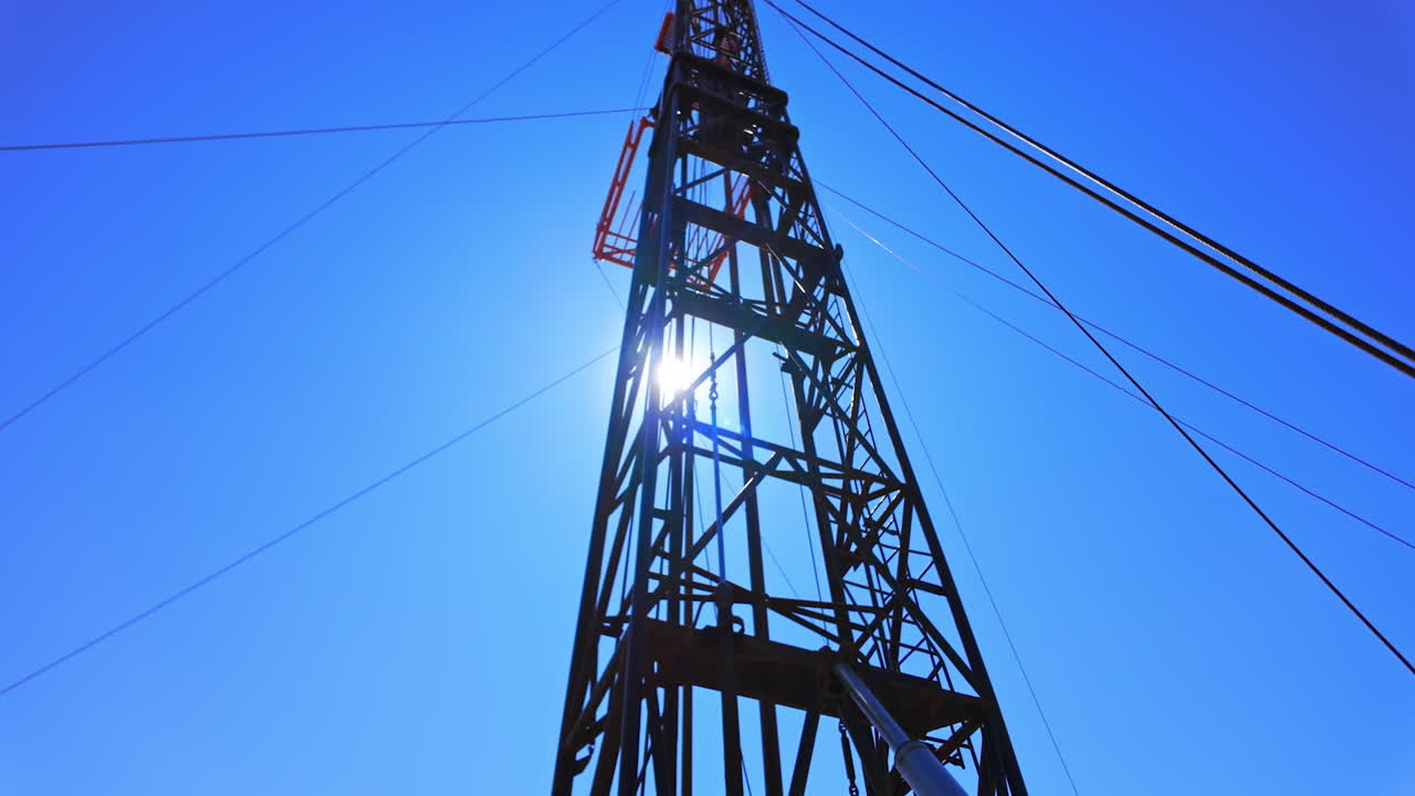 Derrick for drilling oil against the blue sky and bright sun. Low angle view on the rig tower with metal wires attached.