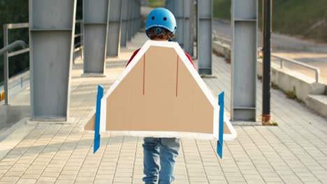 Vista-Trasera-De-Un-Niño-Pequeño-Con-Casco-Y-Suéter-Rojo-Con-Alas-De-Avión-De-Cartón-Caminando-Por-El-Puente-Y-Jugando-Como-Piloto