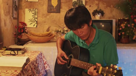 young asian male playing a guitar while on a chair in a rustic room