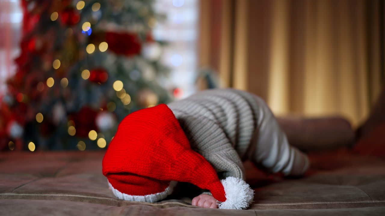 Adorable Caucasian baby with big blue eyes lies looking at camera. Lovely baby boy wearing Santa cap and warm sweater. Blurred Christmas tree at backdrop.