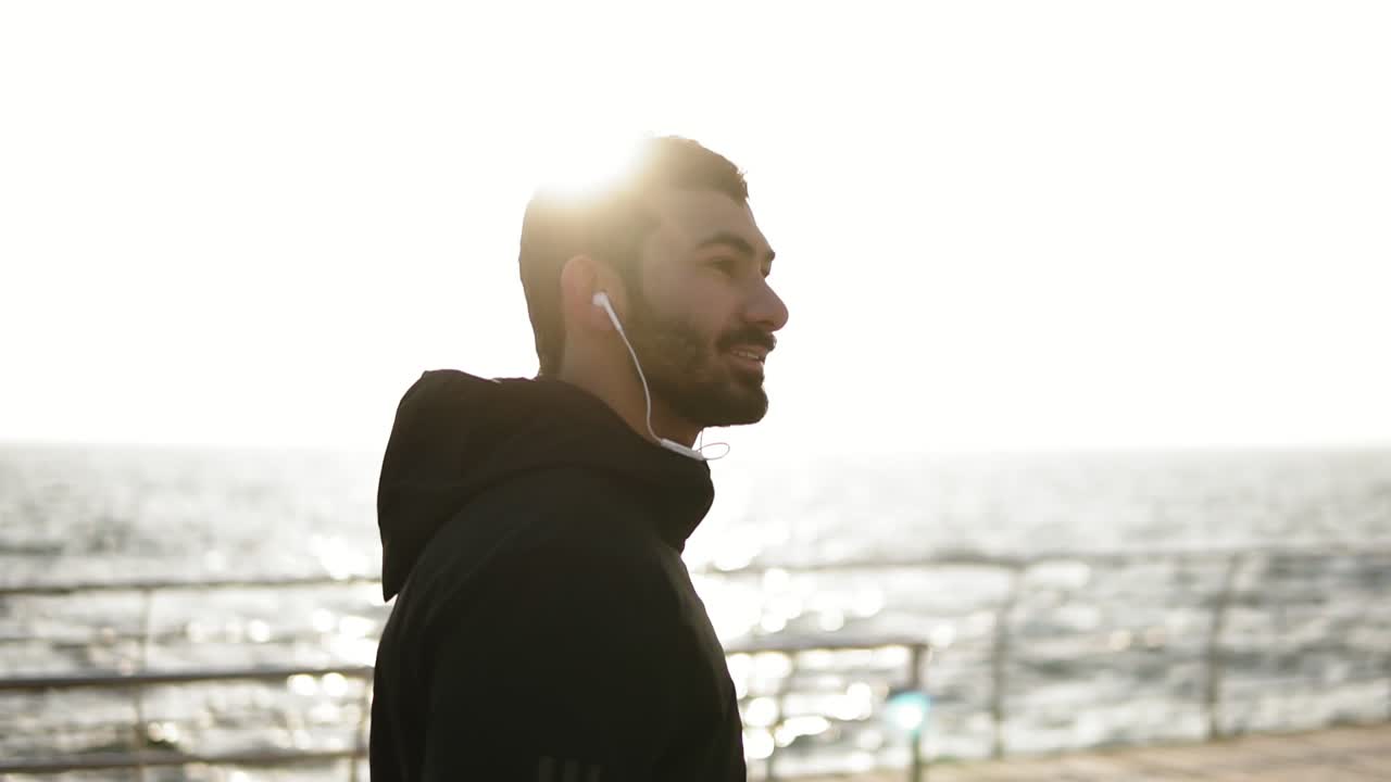 joven hombre atractivo con barba y apariencia caucásica sonriendo y mirando a la cámara mientras camina al aire libre. vista al mar ligeramente reconocible.