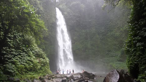 una gran cascada que cae sobre grandes rocas negras en la jungla de bali con un grupo de turistas de pie y observándola