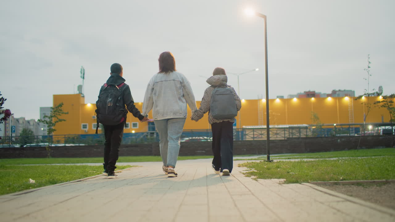 mum holding hands with two children walking down urban park path at dusk capturing family bonding support during evening stroll in green public space near residential area