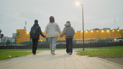mum holding hands with two children walking down urban park path at dusk capturing family bonding support during evening stroll in green public space near residential area