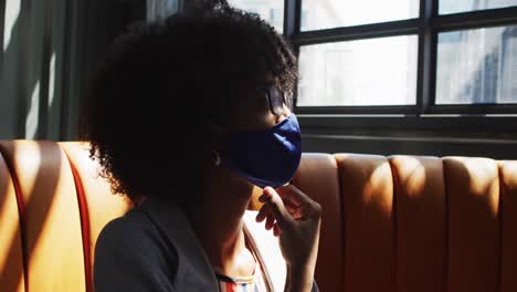 african american woman wearing face masks sitting in cafe looking at window