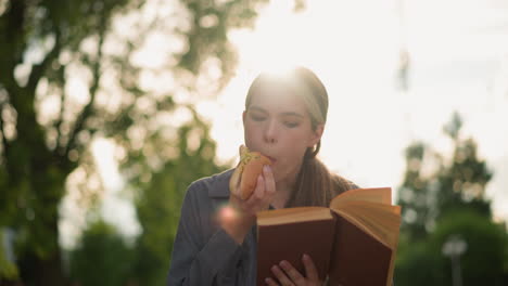 dama comiendo un bocadillo mientras lee un libro al aire libre, sentada en un banco del parque rodeada de vegetación, con árboles borrosos y rayos de luz solar que se reflejan suavemente en el fondo