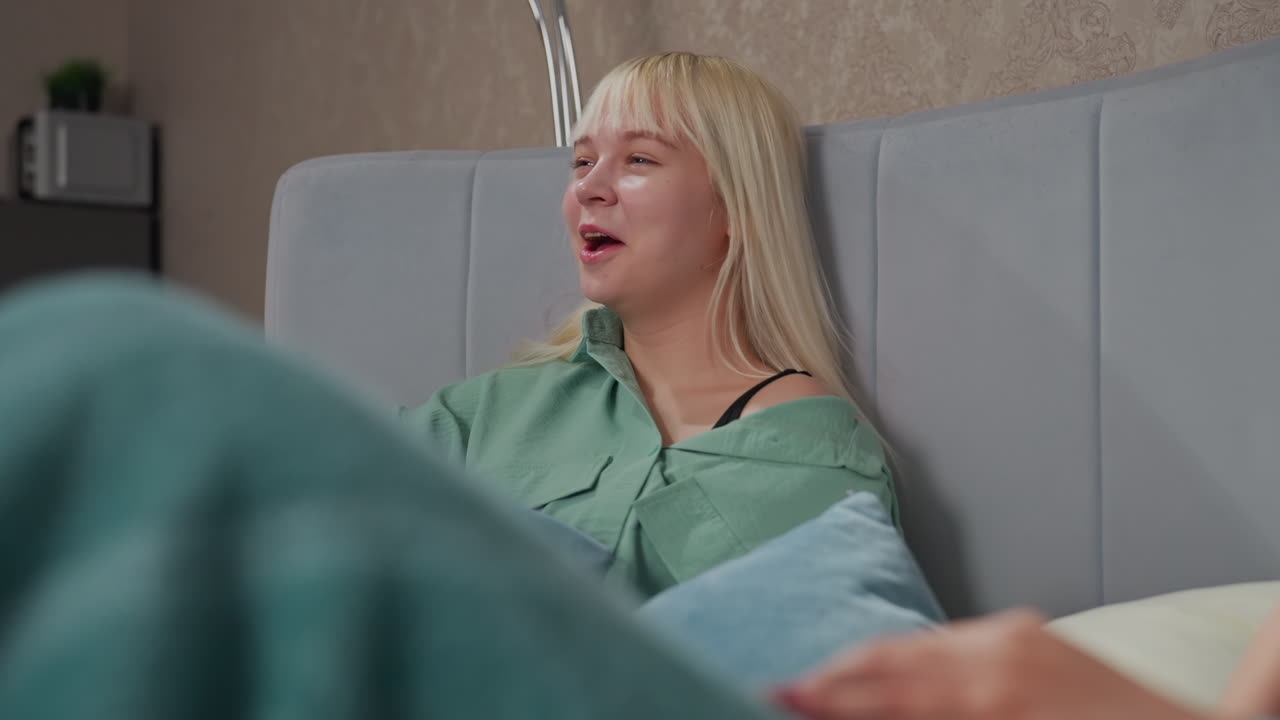 Happy blonde girl laughing joyfully while lying on bed with friend partially visible under blanket, cozy bedroom setting with soft lighting, decorative wallpaper, and casual relaxed mood