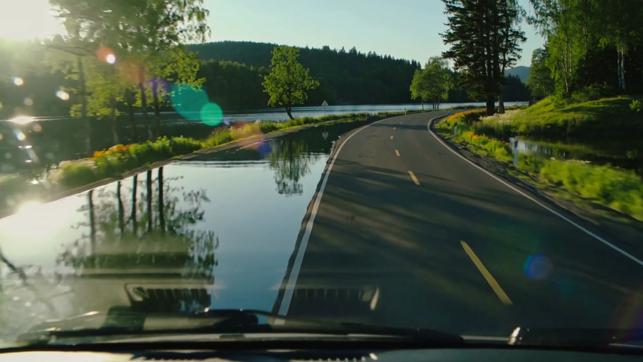 Starting vehicle windshield view tracking two-lane road curving right along lake, with lens flare