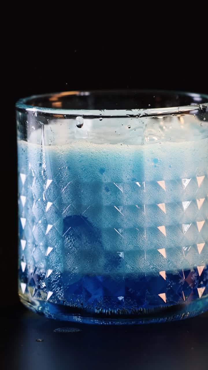 Primo piano verticale di un liquido blu intenso che si versa in un bicchiere lavorato con cubetti di ghiaccio
