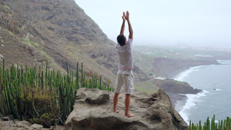 Amidst-mountains-and-overlooking-the-ocean,-the-young-man-seamlessly-merges-meditation-and-fitness,-engaging-in-sun-salutation-yoga-exercises