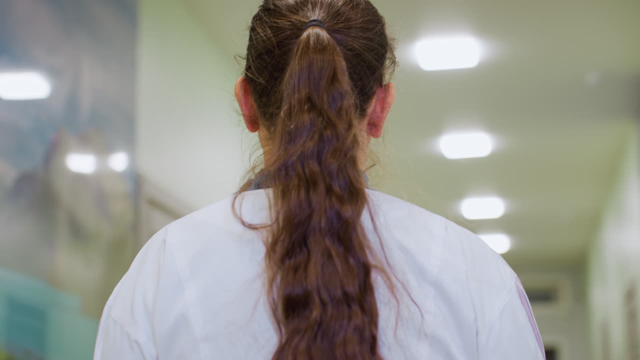 Healthcare assistant with long brown ponytail walks through bright hospital lobby corridor under ceiling lights wearing white coat focused on destination as warm lighting fills clinical hallway space
