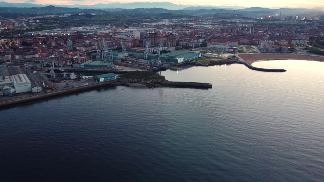 Vista aerea della costa della città di Gijón nel nord della Spagna al tramonto, spiaggia di sabbia e porto commerciale.