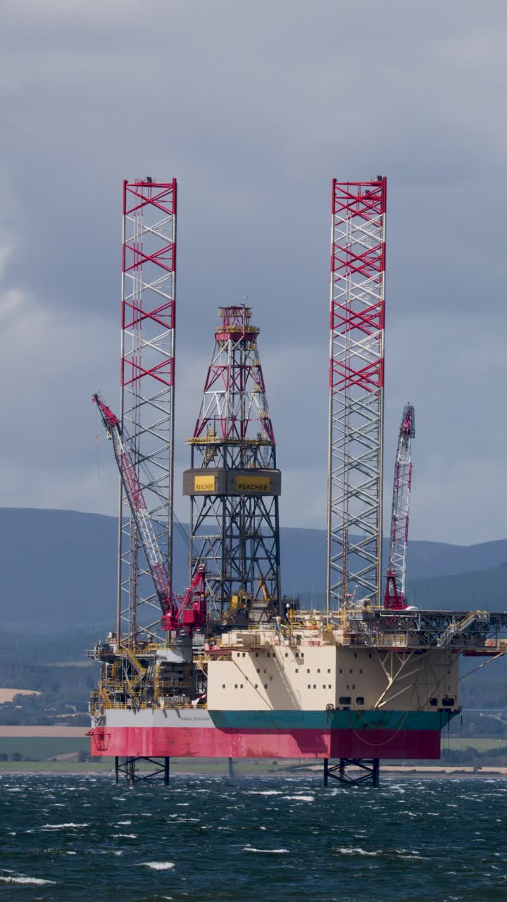 Offshore olieplatform med kraner i drift, overskyet dagslys, stabilt kamera, skotsk højlandskystlinje synlig.