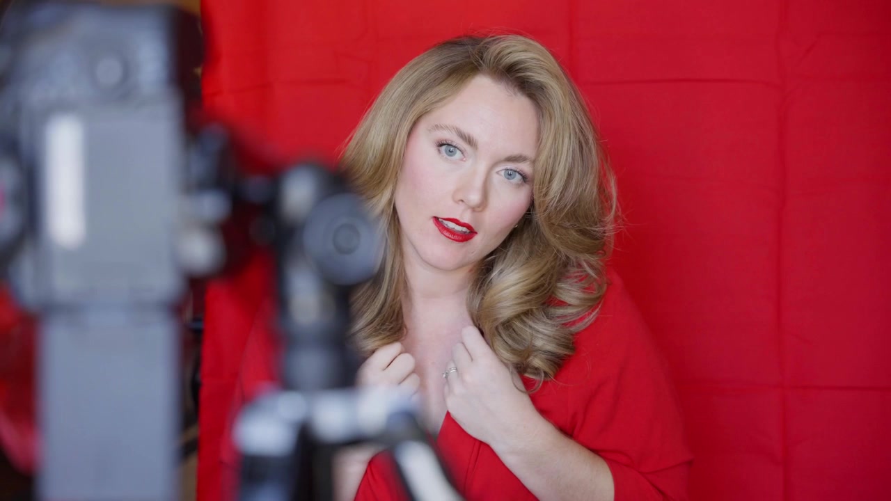 Blonde woman in a red outfit posing during an indoor fashion shoot, with camera equipment in foreground, red backdrop and polished beauty styling.