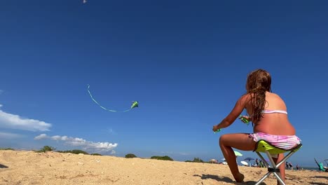 red-haired little girl sitting on beach chair enjoys flying a kite on hot summer day