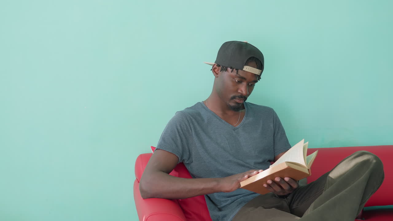 Jeune homme concentré en tenue décontractée, assis sur un canapé rouge avec une jambe relevée, feuilletant les pages d’un livre qu’il tient entre les mains, devant un mur turquoise, semblant absorbé par une activité de lecture attentive.