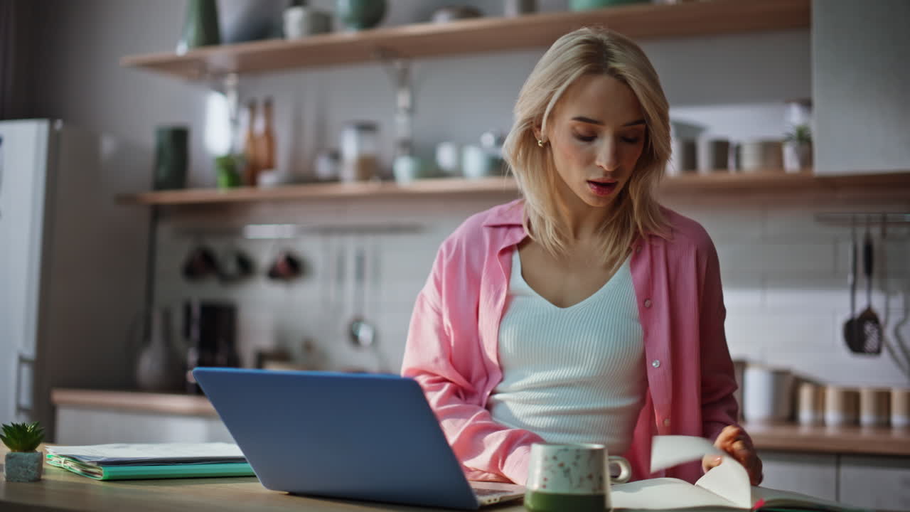 Intelligenter Freelancer macht sich Notizen auf einem Notizblock und schaut auf den Laptop-Bildschirm in der Küche (Nahaufnahme)