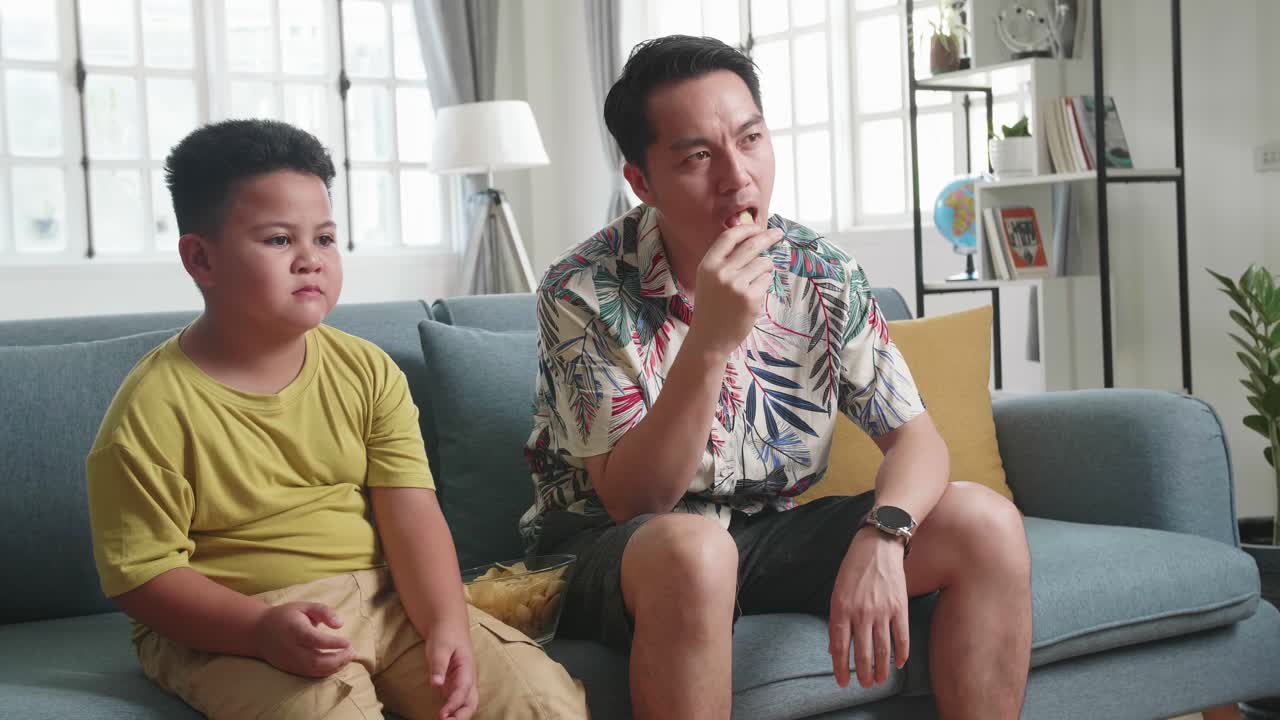 Asian Father And His Son Are Watching Tv While Eating Potato Chips At Home