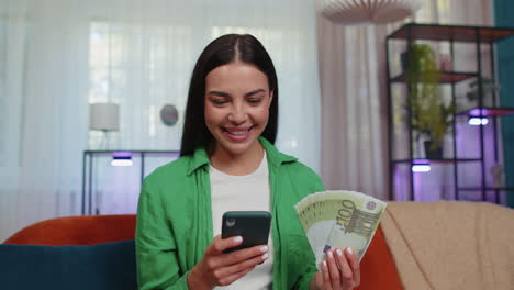 Smiling-happy-young-woman-at-home-counting-money-cash-use-smartphone,-income,-saves,-lottery-win