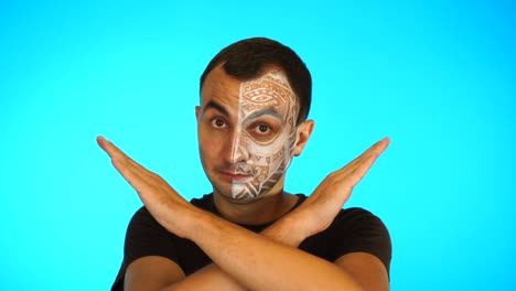 man with painted face is crossed arms at camera against blue background