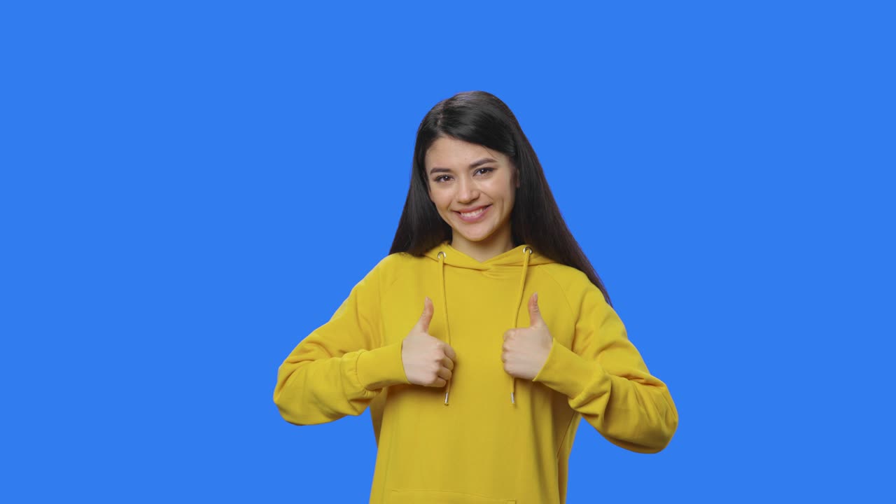 retrato de una bonita morena señalando los dedos y mostrando el pulgar como. mujer joven con cabello largo en sudadera amarilla posando en el estudio con fondo de pantalla azul. de cerca. cámara lenta lista 59.94fps