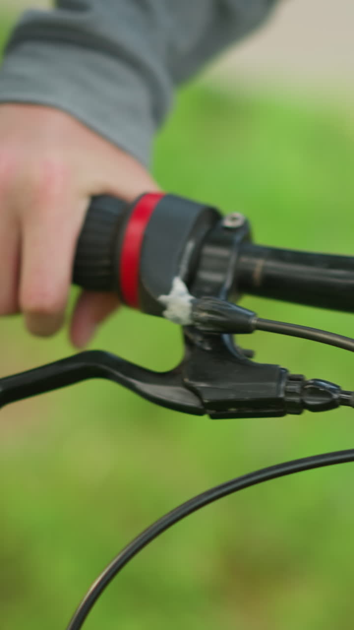 primer plano de un individuo con pantalones negros y tela gris, se acerca a una bicicleta estacionada, ajustando la palanca del freno en un campo cubierto de hierba, la persona sostiene el manillar con un agarre firme