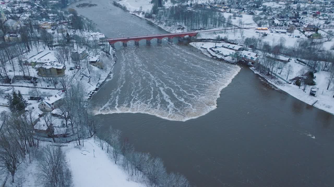 vista aérea de los rápidos del río venta durante la inundación de invierno, viejo puente de ladrillo rojo, kuldiga, letonia, día de invierno nublado, amplia toma de drone moviéndose hacia adelante inclinándose hacia abajo