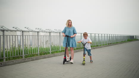 a mother and her son ride scooters side by side on a paved walkway, with iron rails bordering the path, they enjoy a peaceful outdoor activity together