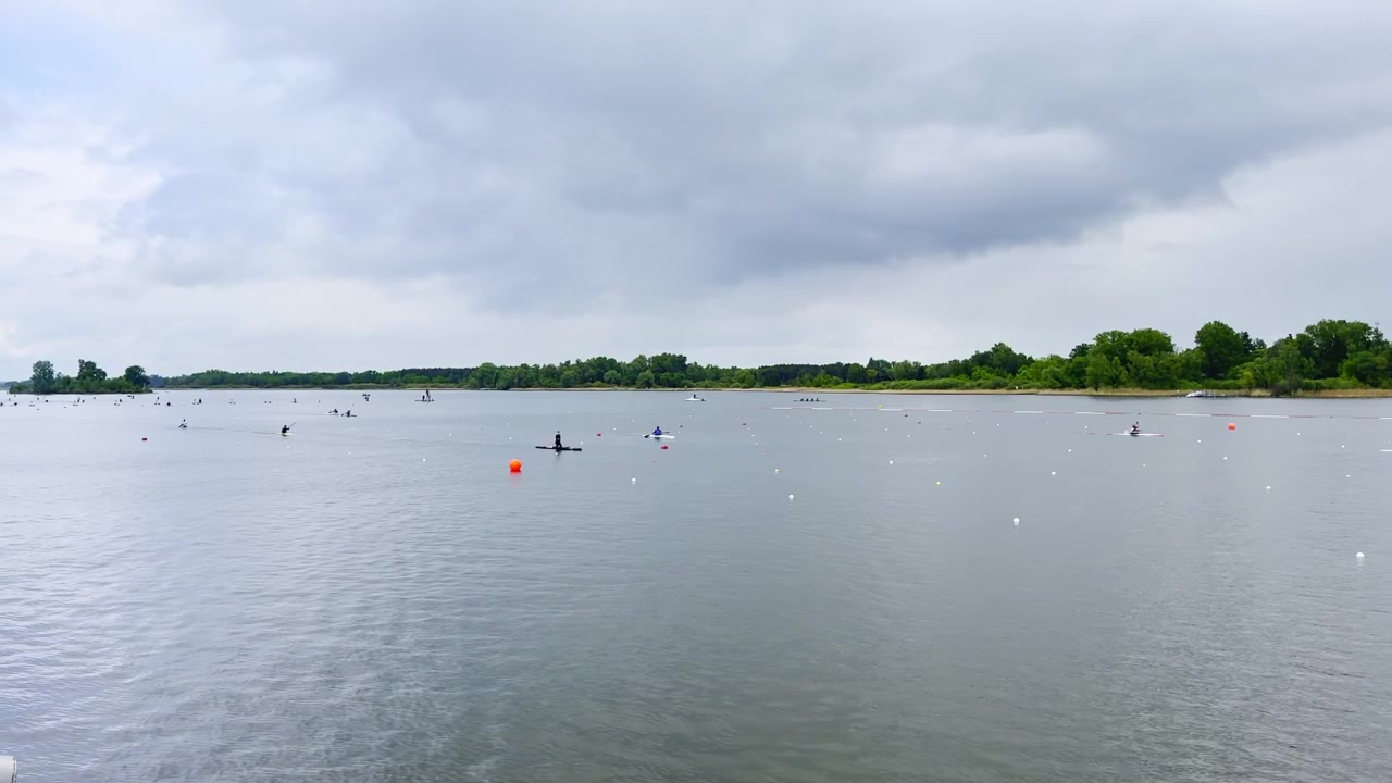 Wide view of paddlers training on a Brandenburg canoe sprint lake, with scattered lane buoys, calm grey water, shoreline trees and cloudy race-course atmosphere.