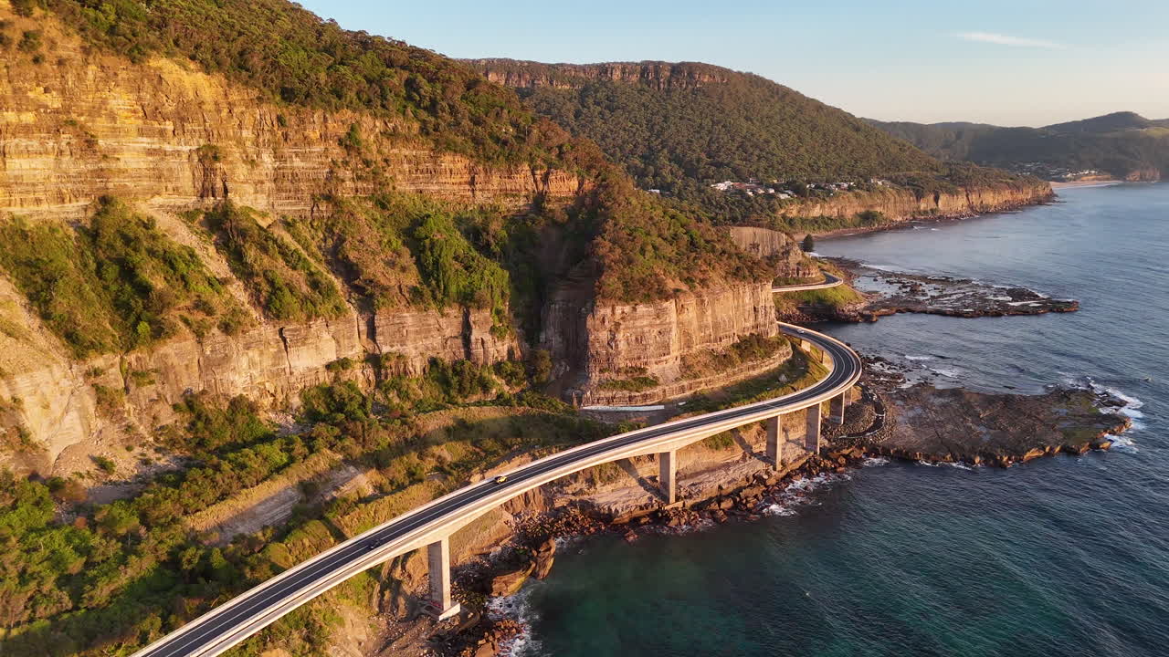 Wide aerial shot of a car on the Sea Cliff Bridge, highlighting the dramatic coastline and winding roadway. Tracking drone footage.