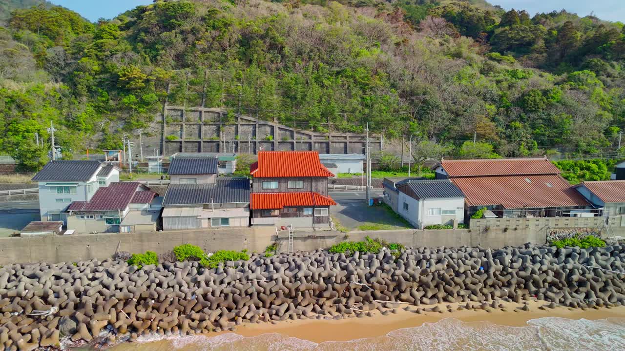 An aerial shot pans left, following the coastline of a quaint Japanese village protected by a large tetrapod sea wall, with lush green hills behind