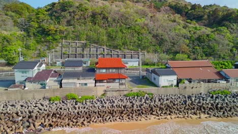 an aerial shot pans left, following the coastline of a quaint japanese village protected by a large tetrapod sea wall, with lush green hills behind