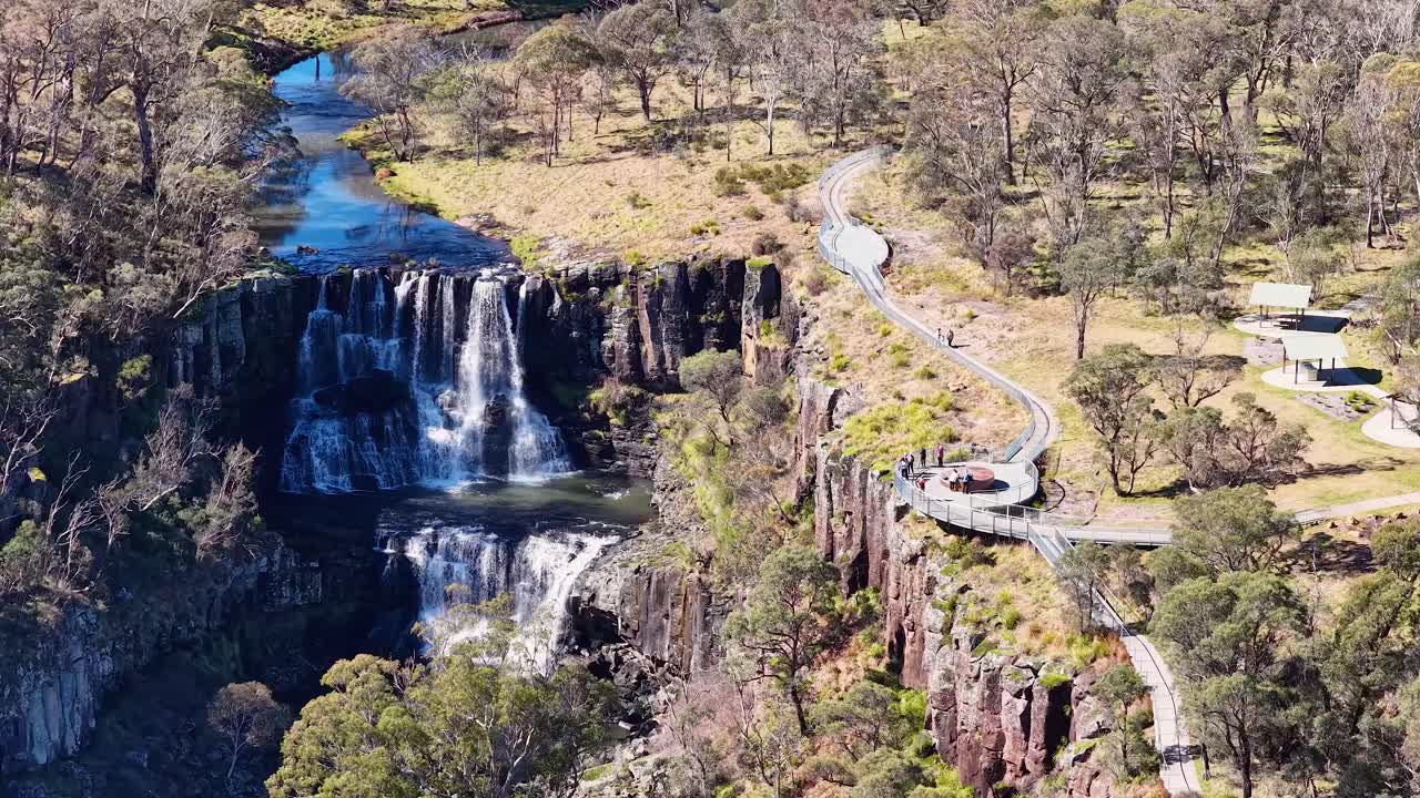 Drone footage glides toward a clifftop viewing platform overlooking a dramatic multi-tiered waterfall, surrounded by rugged bushland in bright daylight