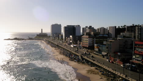 Luftaufnahme-Des-Strandes-Farol-Da-Barra-Mit-Den-Menschen,-Die-Den-Sonnigen-Tag-Genießen,-Und-Den-Gebäuden-Im-Hintergrund,-Salvador,-Bahia,-Brasilien