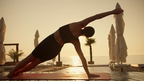 Rear-view-of-a-brunette-girl-in-a-black-sports-summer-uniform-doing-plank-exercises-on-a-red-mat-on-the-Sunny-seashore-during