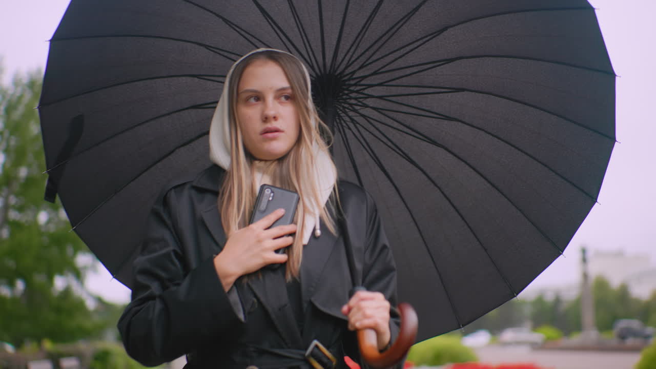 Gentlelady in black coat with hood holds umbrella and talks on phone outdoors during rainy day, walking on city street with blurred greenery and buildings in background, appearing serious and focused