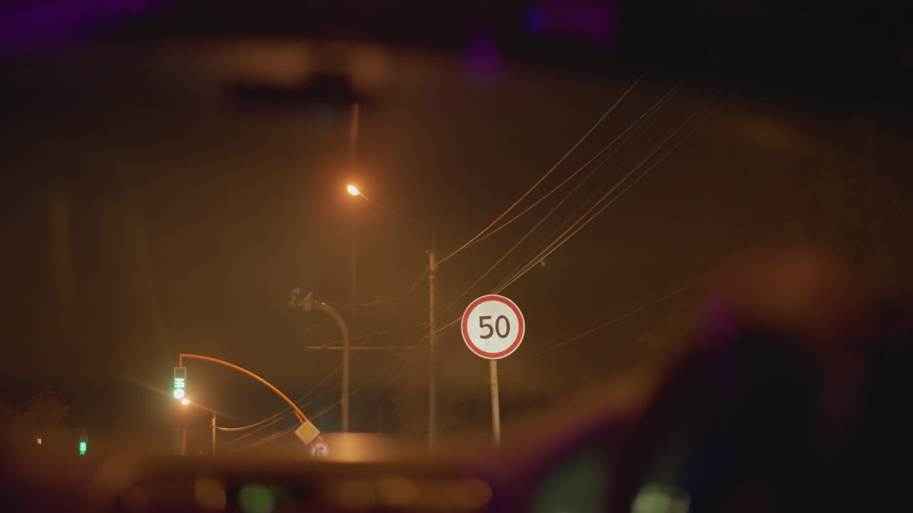 Conducción nocturna bajo la lluvia a través del parabrisas, farolas brillantes, señal borrosa, gotas de lluvia resbalando por el cristal, siluetas ámbar en el salpicadero, taxista mirando por el espejo, mensajero nocturno esperando en el semáforo en rojo