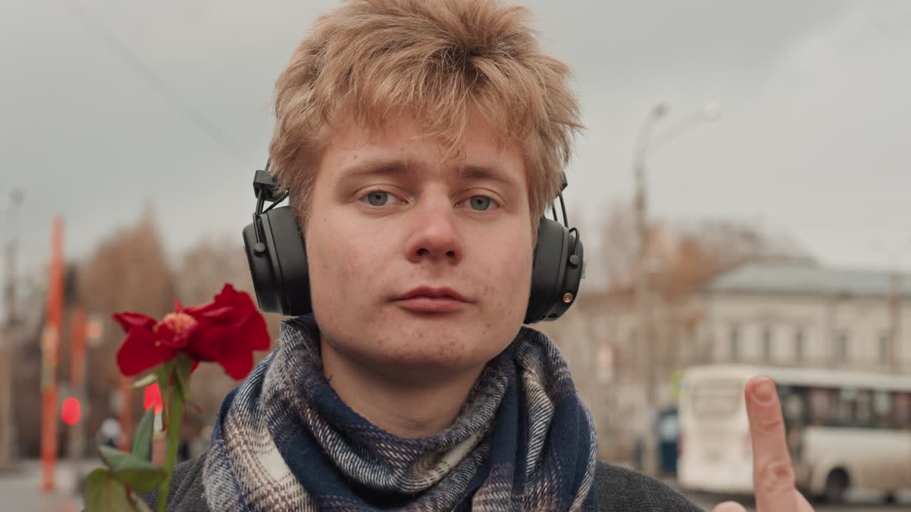 Creatore bianco che ascolta a occhi chiusi con le cuffie, atmosfera da cantautore vicino a una fermata dell'autobus urbana con una rosa a lato, artista con sciarpa meditativo nella luce autunnale, ritratto stradale calmo e riflessivo.