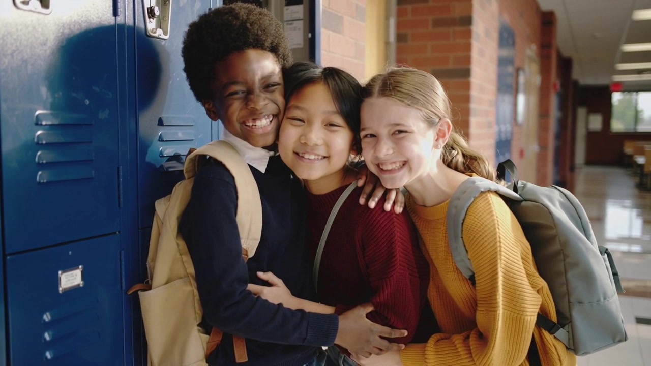 Trio sorridente con zaini che si stringe mentre la telecamera zooma in un corridoio scolastico vicino agli armadietti blu, creando legami.