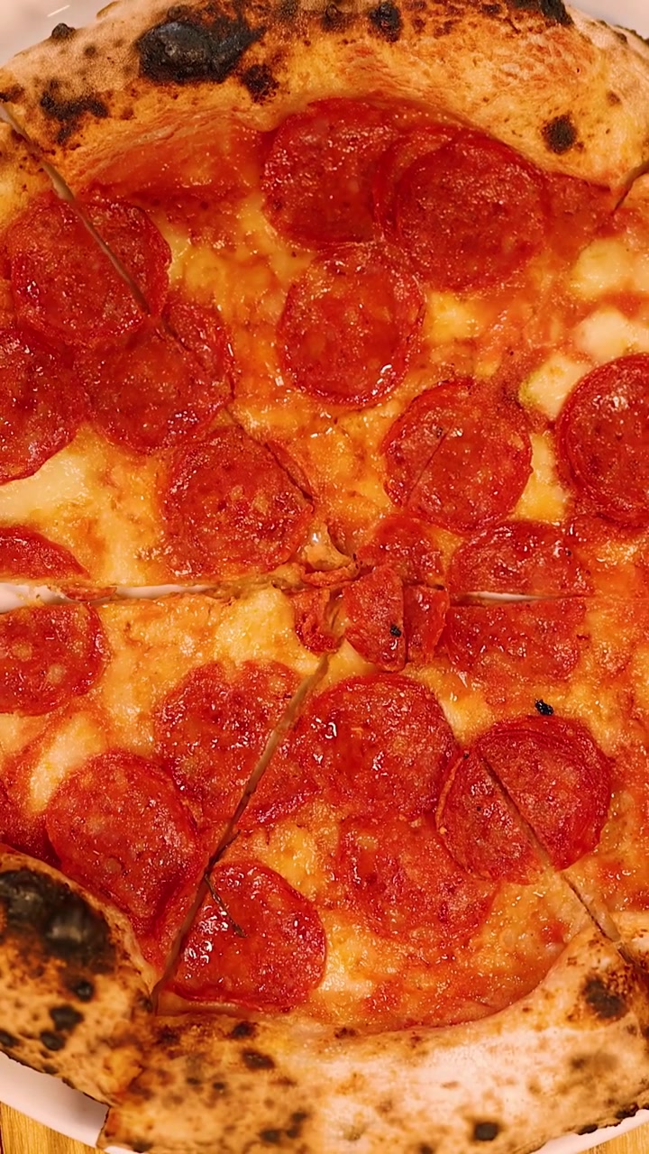 Inquadratura dall'alto di mani che tagliano una pizza al lievito madre e salame piccante su un tavolo di legno di un ristorante.