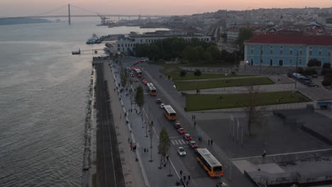 Aerial-reveal-of-busy-car-traffic-on-the-coast-of-Lisbon,-Portugal-with-iconic-red-bridge-Ponte-25-de-Abril-in-the-background