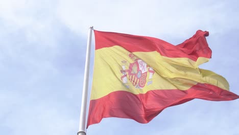 Toma-Detallada-De-La-Bandera-Española-Ondeando-En-El-Viento-En-Cámara-Lenta-Con-Cielo-Azul-Y-Nubes-En-El-Fondo